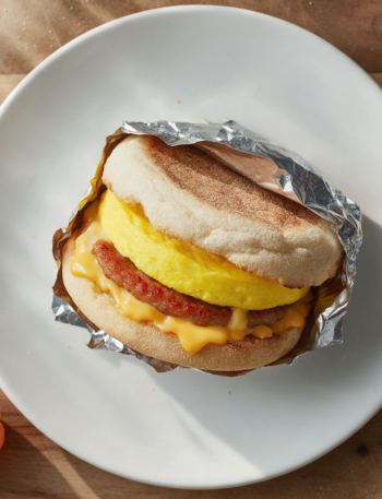 Sausage egg cheddar sandwich on a toasted muffin, cheese melting, foil peeled back on a rustic wood board in morning light