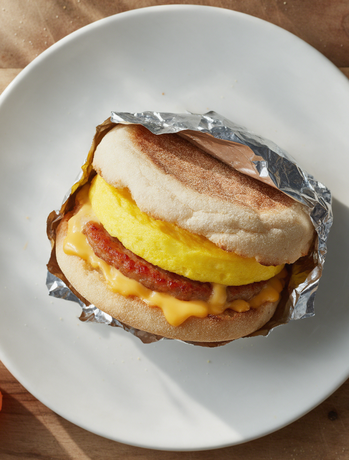 Sausage egg cheddar sandwich on a toasted muffin, cheese melting, foil peeled back on a rustic wood board in morning light
