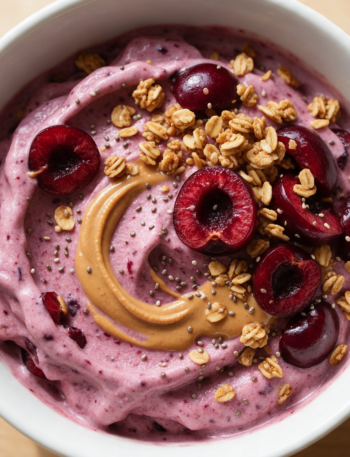 Overhead shot of a cherry Greek yogurt smoothie bowl topped with granola, chia seeds, and an almond butter swirl on wood.