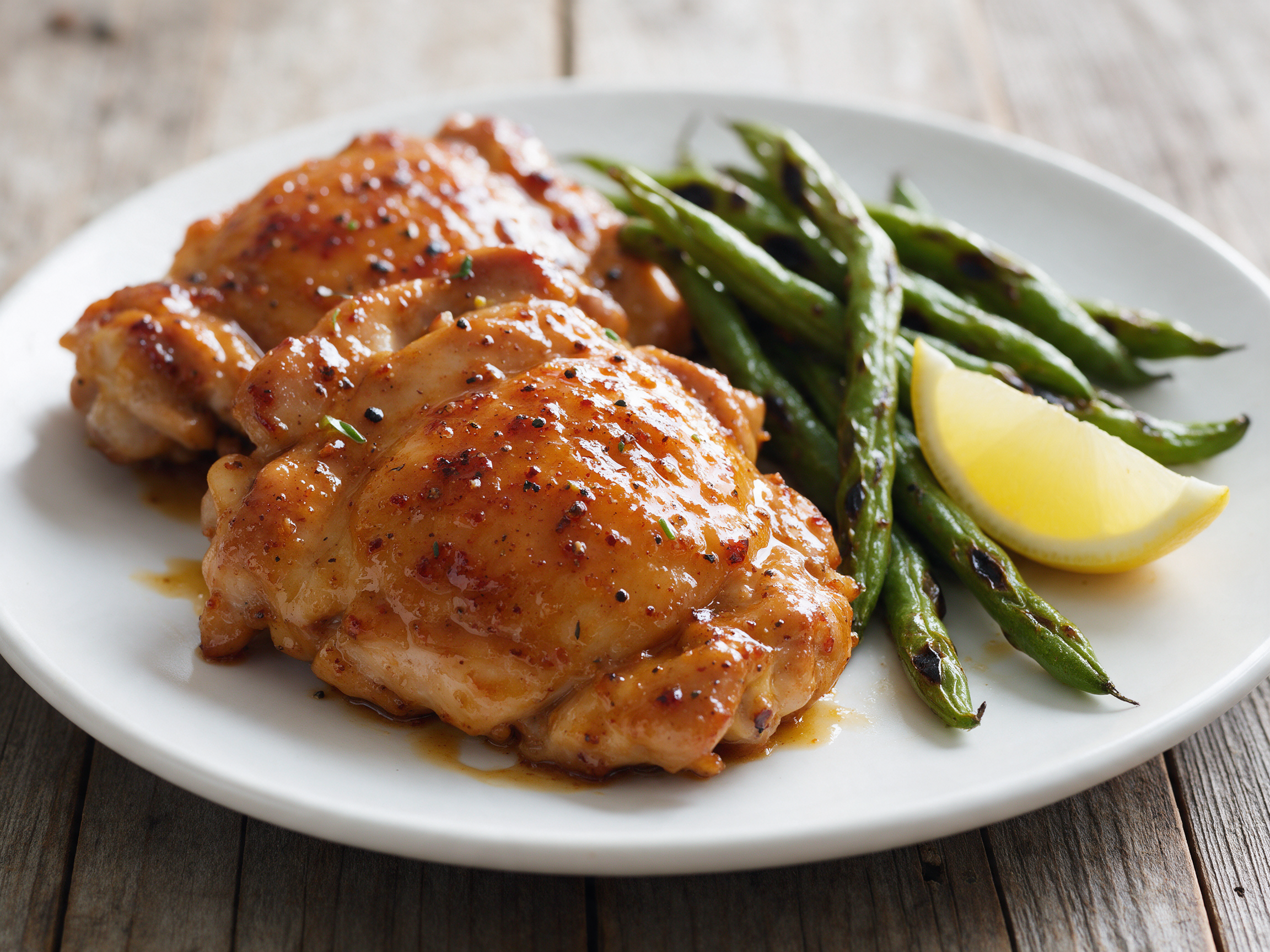 Glazed honey mustard chicken thighs beside blistered green beans on a white plate, shot at 45 degrees on a wooden table